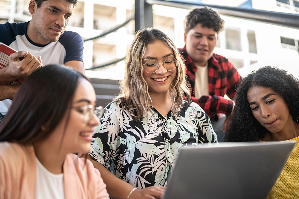 Group of diverse university students sitting on outdoor stairs, with a young woman smiling and showing something on her laptop to her friends