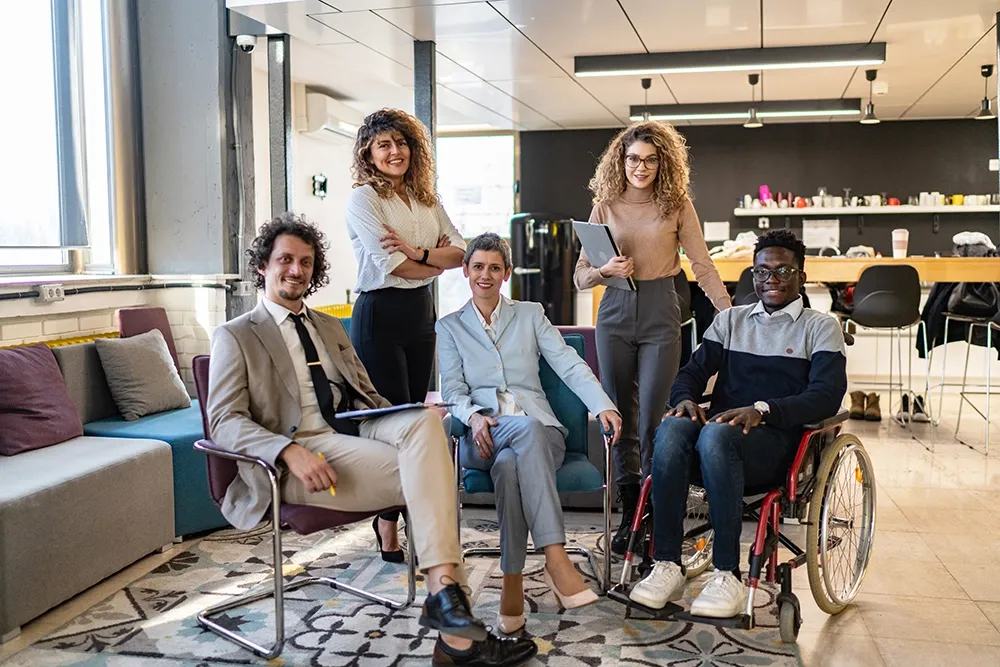 Group of diverse business professionals, including a man in a wheelchair, having a discussion in a modern office space, representing inclusion and accessibility